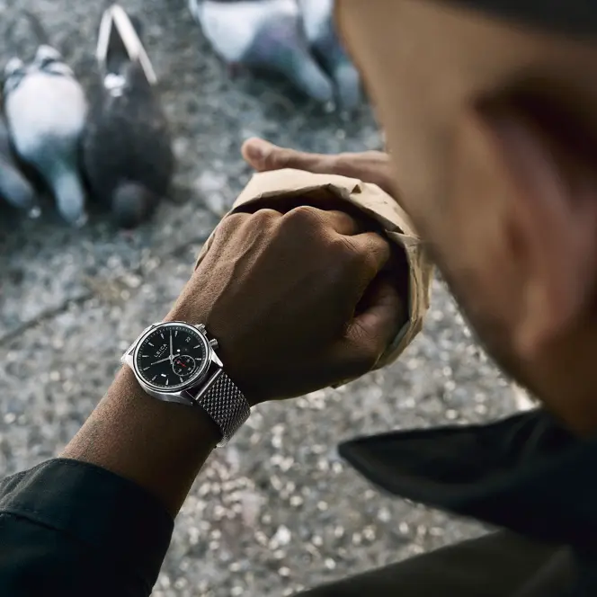Jason Roman checks his Leica Watch while feeding pigeons in an urban public space, capturing a candid moment of everyday city life and human interaction with wildlife.