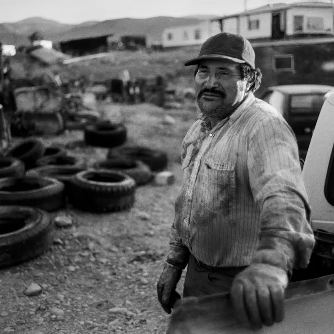 A man in a worn work shirt leans against a pickup truck in a rural scrapyard in Montana. Old tires, machinery, and trailers fill the foreground, with the Rocky Mountains in the distance.