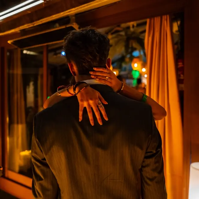 Documentary photograph of a couple embracing at a warmly lit evening celebration, capturing intimacy, human connection, and contemporary social life.