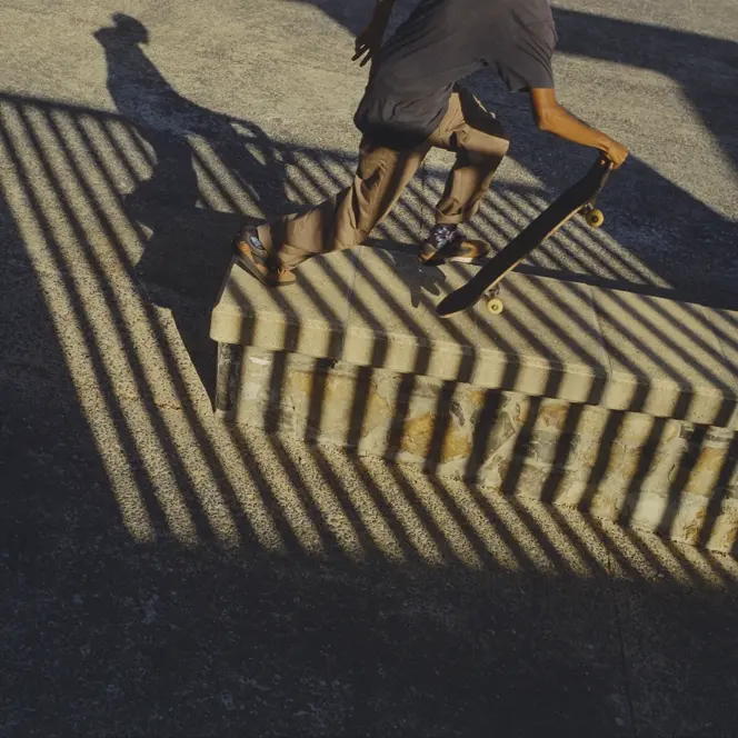 Skateboarder carrying a board through dramatic striped shadows at a skatepark, capturing movement, urban culture and graphic light.