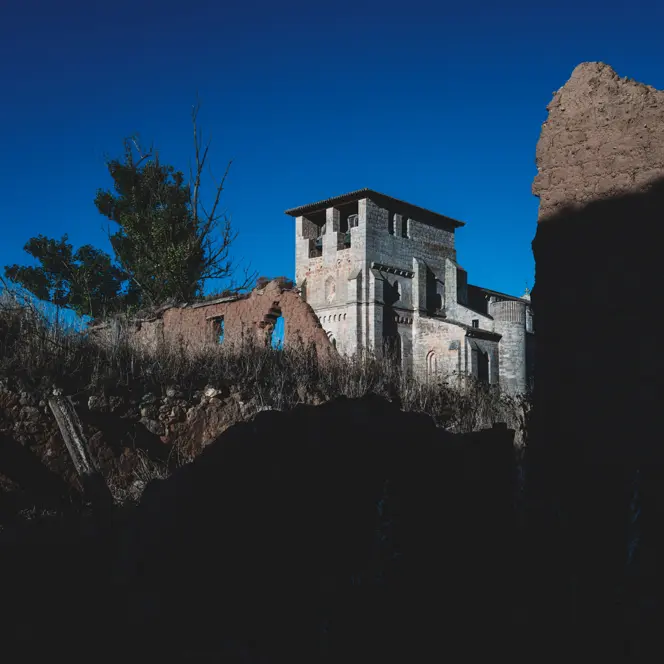 Historic stone ruins and a tower rise from a rugged landscape beneath a deep blue sky, framed by dramatic light and shadow.
