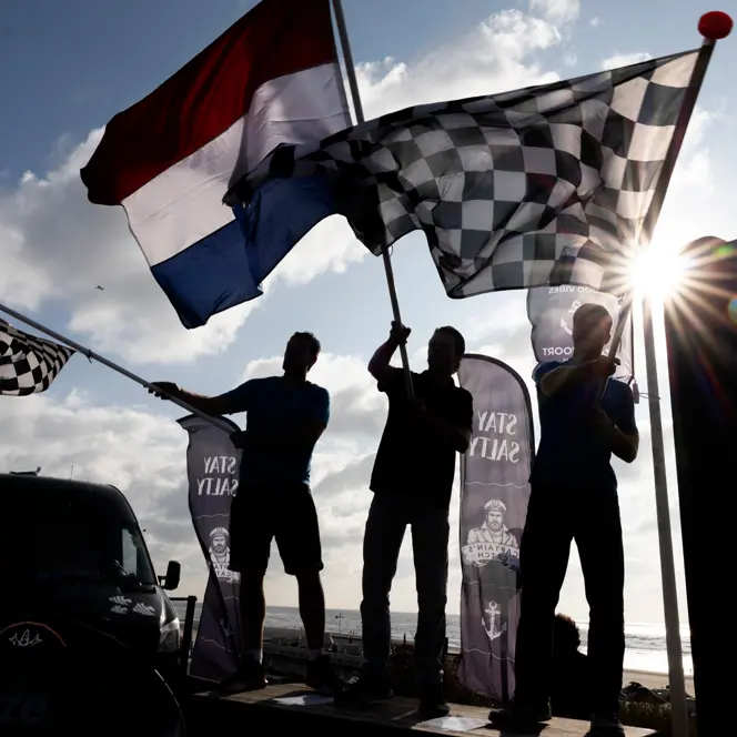 Silhouetted fans wave Dutch and chequered flags at a Formula 1 event in Zandvoort, with bright sunlight breaking through above the seaside circuit.