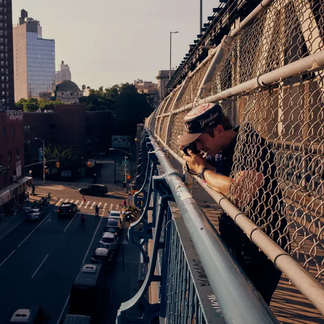 Paul Hepper leaning through the fence of a New York City bridge, taking a street photograph above traffic and urban buildings in warm evening light.