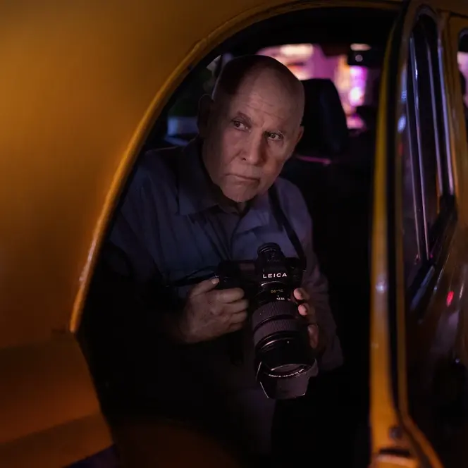 Photographer holding a Leica SL camera while seated inside a yellow taxi at night, illuminated by colorful urban lights.