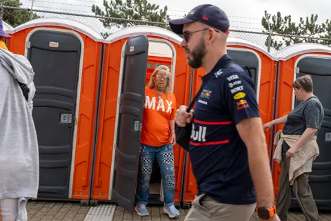 Formula 1 fans at the Dutch Grand Prix in Zandvoort pass a row of bright orange portable toilets, including a woman in orange and a Red Bull supporter.