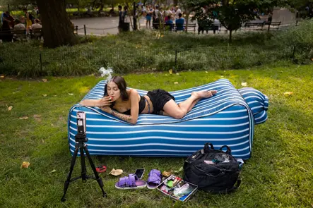 Person relaxing on an inflatable lounge in a city park while livestreaming, capturing contemporary urban life and social media culture.