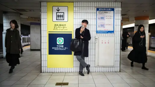 Commuters moving through a Tokyo subway station as a man waits beneath wayfinding signs. Urban documentary photography highlighting public transport, city infrastructure, and daily routines in Japan.