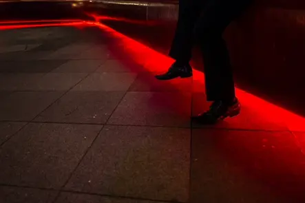 Documentary photograph of a person sitting beneath striking red neon lighting, capturing the atmosphere of a nighttime event with dramatic light and shadow.