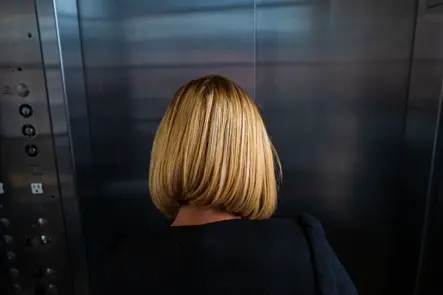 Documentary photograph of a woman seen from behind inside an elevator, highlighting solitude, urban architecture, and everyday life through minimalist composition.