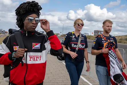 Formula 1 fans in Ferrari and Red Bull team apparel walk toward the Dutch Grand Prix in Zandvoort under a bright, cloudy sky.