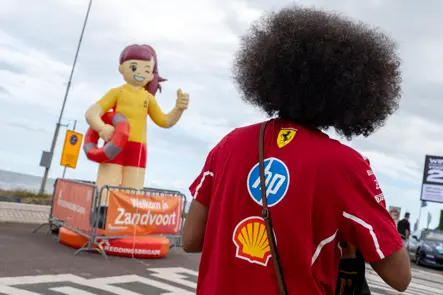 Ferrari fan with a large afro wig at the entrance to the Dutch Grand Prix in Zandvoort, in front of a lifeguard figure and event signage.