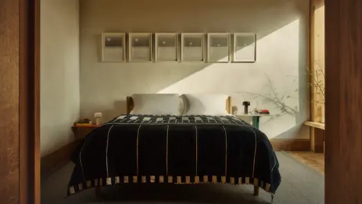A minimalist bedroom with a double bed covered in a black patterned blanket, six framed photographs above the headboard, and warm sunlight falling across the wall.