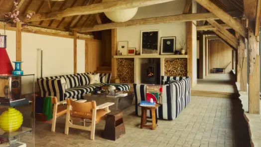 A spacious living room with exposed timber beams, striped sofas, a lit fireplace, stacked firewood, artworks and colorful decorative objects.