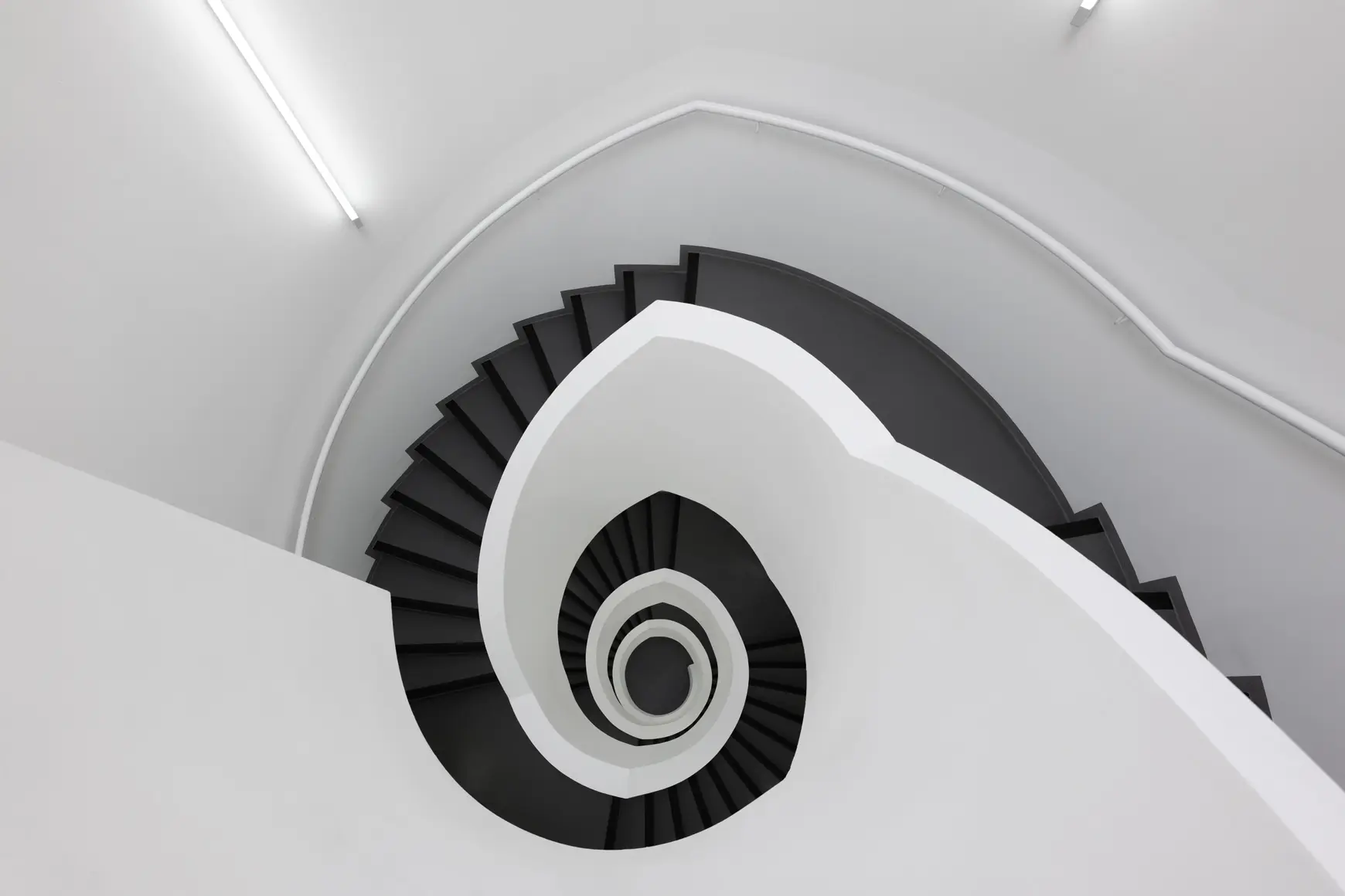 Stairwell looking like a snail shell with white walls and blacks stairs photographed from the top. 