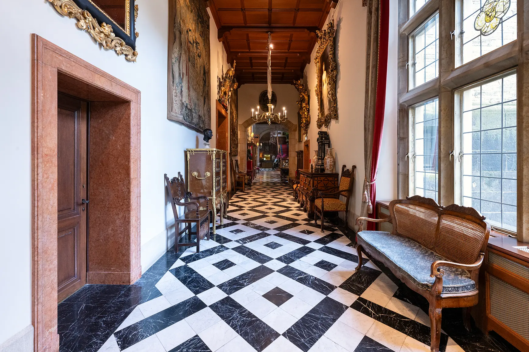 A high and wide hallway with black and white tiles. Window- and doorframes out of wood.