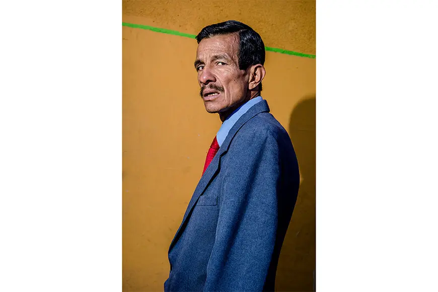 Man with moustache and blue suit in front of yellow wall