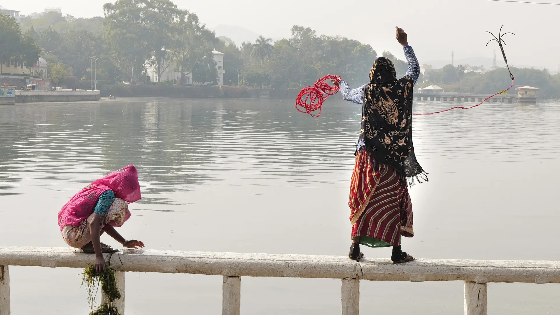 A woman and a child are standing on a railing in front of a lake and are fishing.
