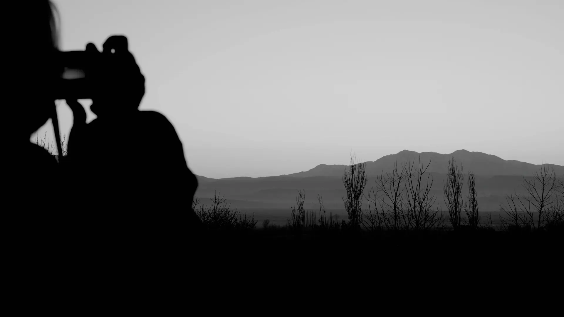 Black-and-white photograph of a person taking a picture of a lake and mountains in the background.