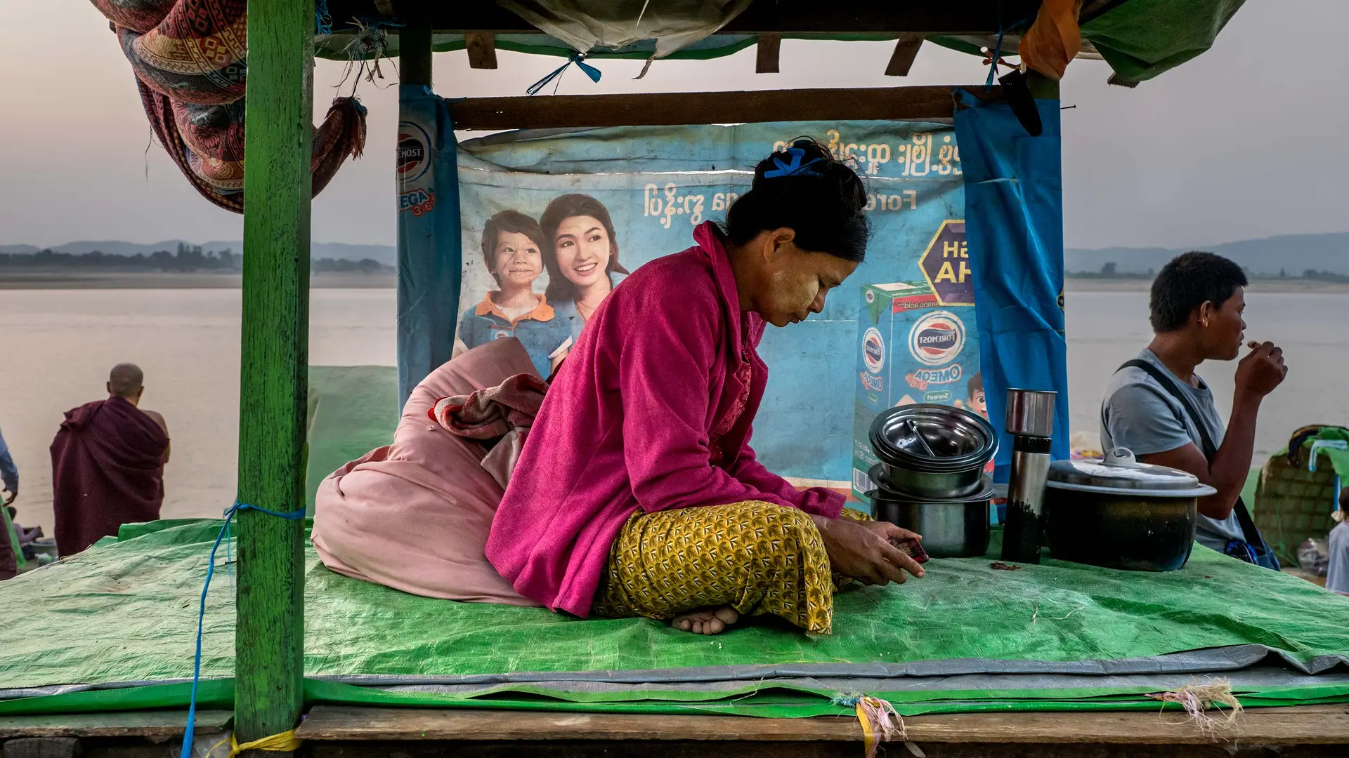 Woman sits on plastic tarp looking at a phone, in background are other people and a river.Woman sits on plastic tarp looking at a phone, in background are other people and a river.