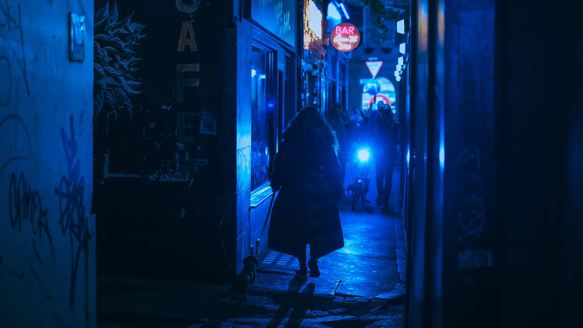 Person walking a dog through a narrow alley at night, illuminated by blue neon lights.