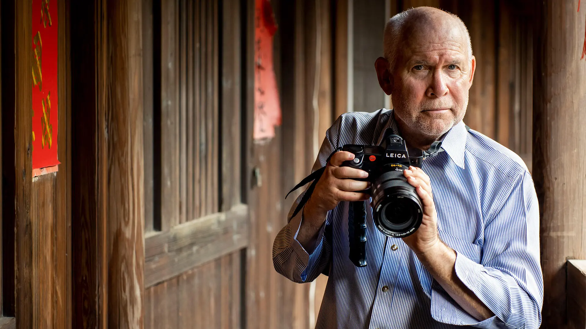Steve McCurry holds a Leica camera and stands in front of a wooden house.