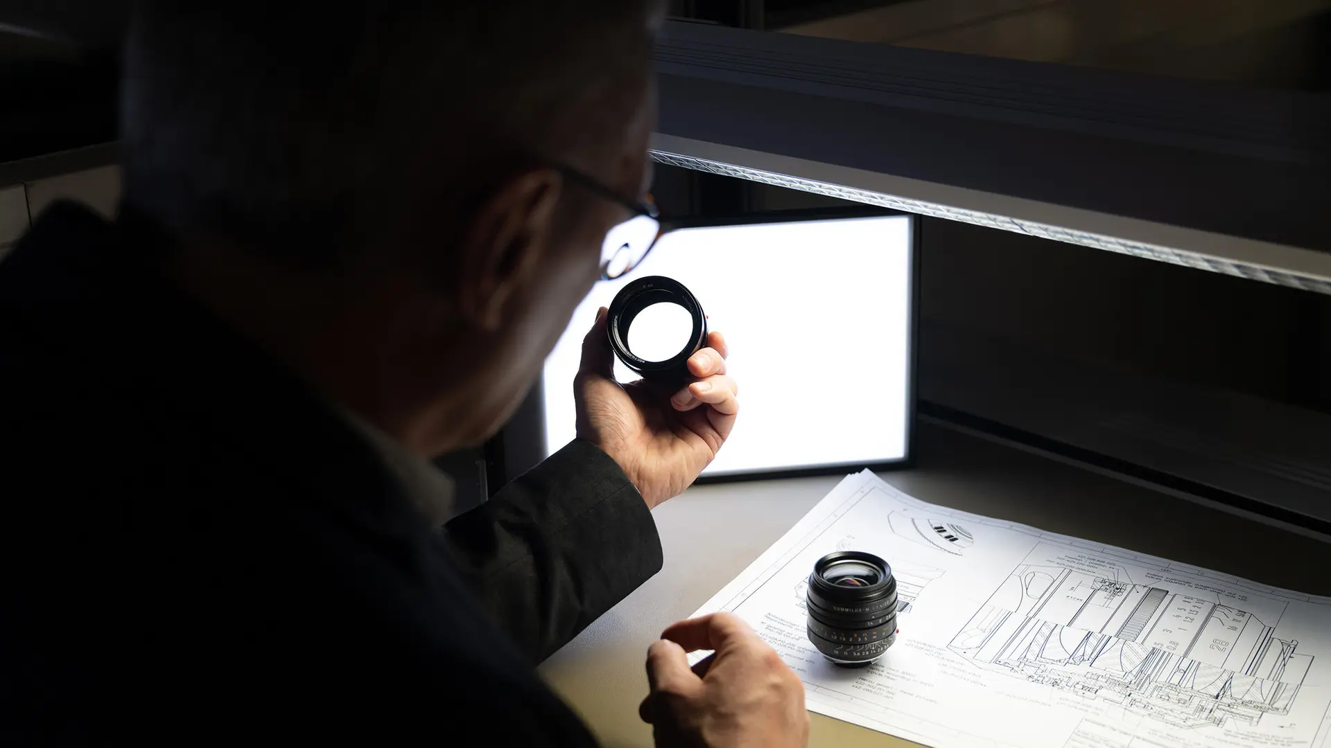 A man inspects a camera lens, technical drawings lie on the table.
