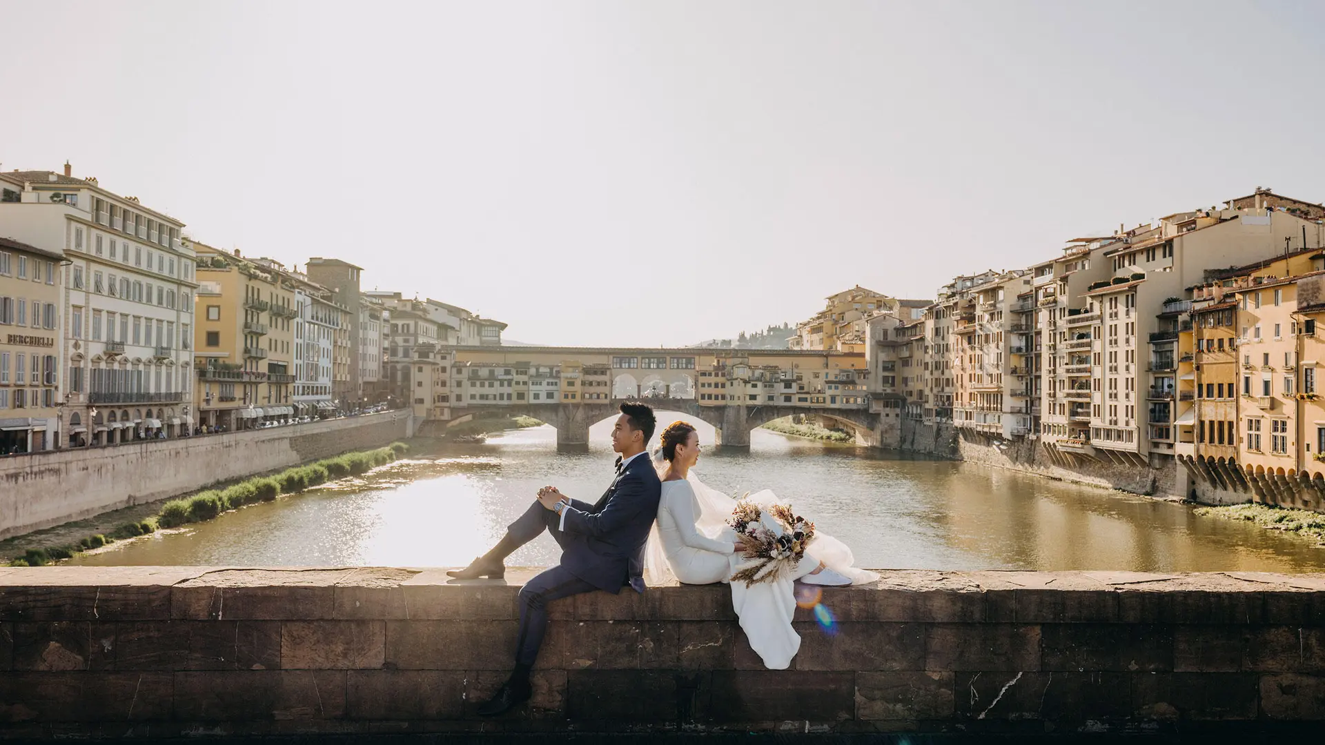 A wedding couple sits on a wall overlooking the river, surrounded by old buildings.