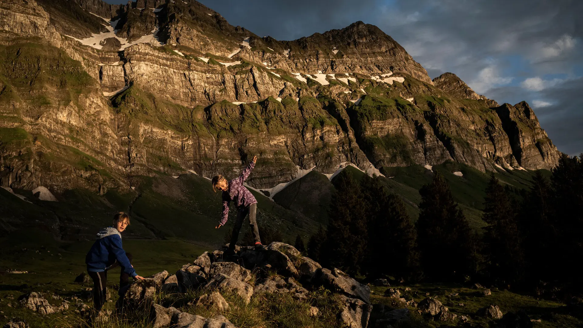 Two children climbing rocks in a mountain landscape at sunset.