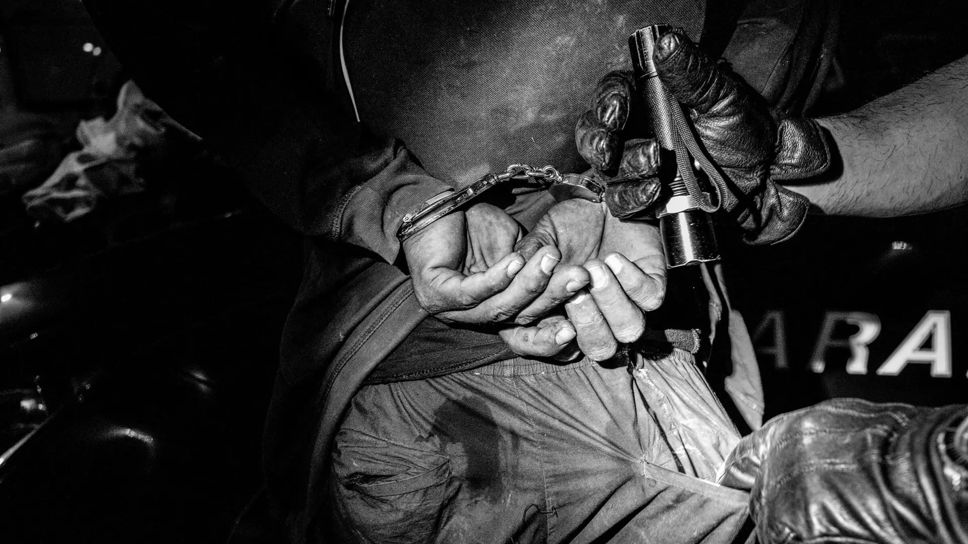 Close-up of a handcuffed person and another person searching his pants pocket with a flashlight.