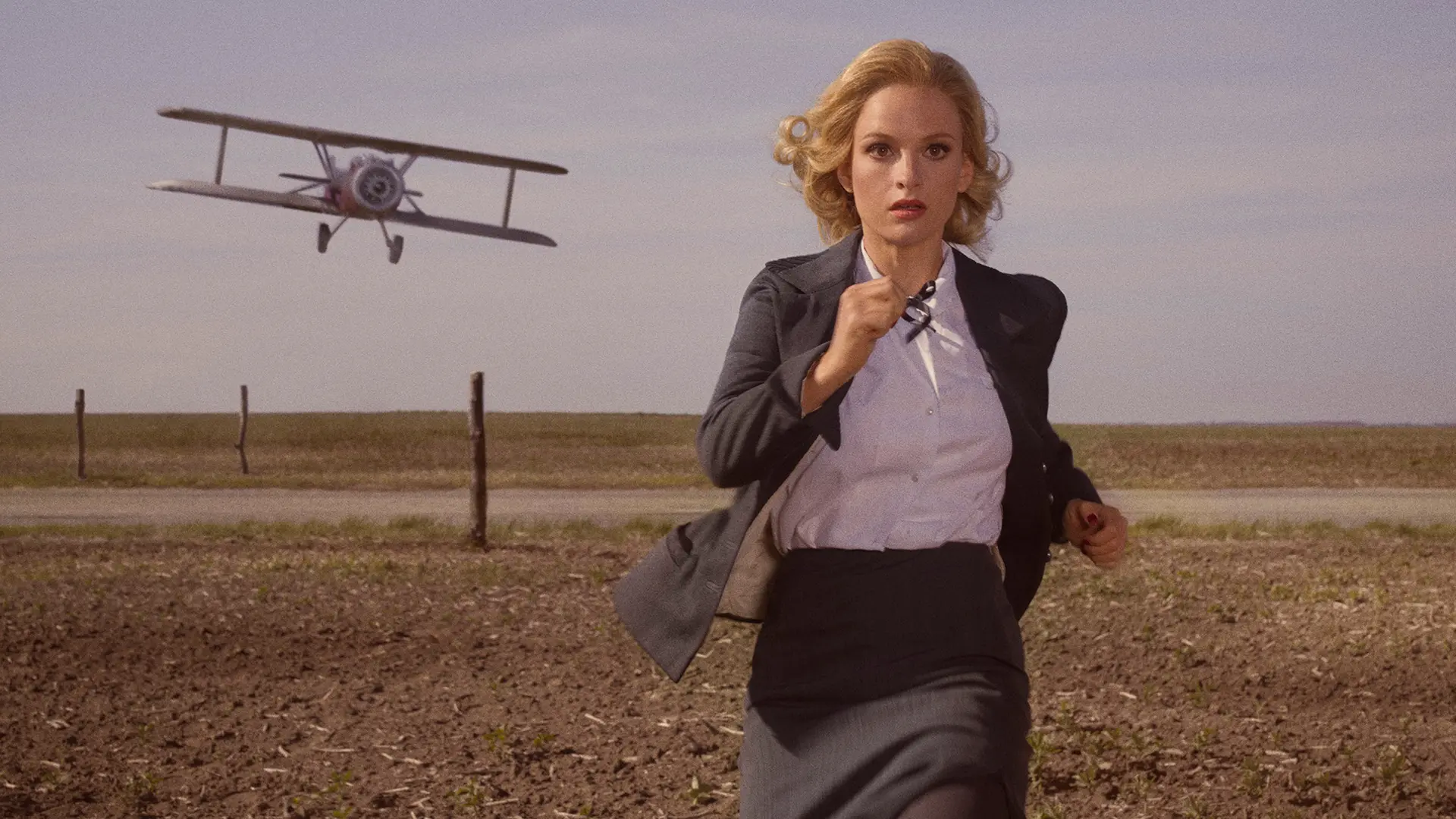 Woman in suit running across a field with an old propeller plane flying behind her.