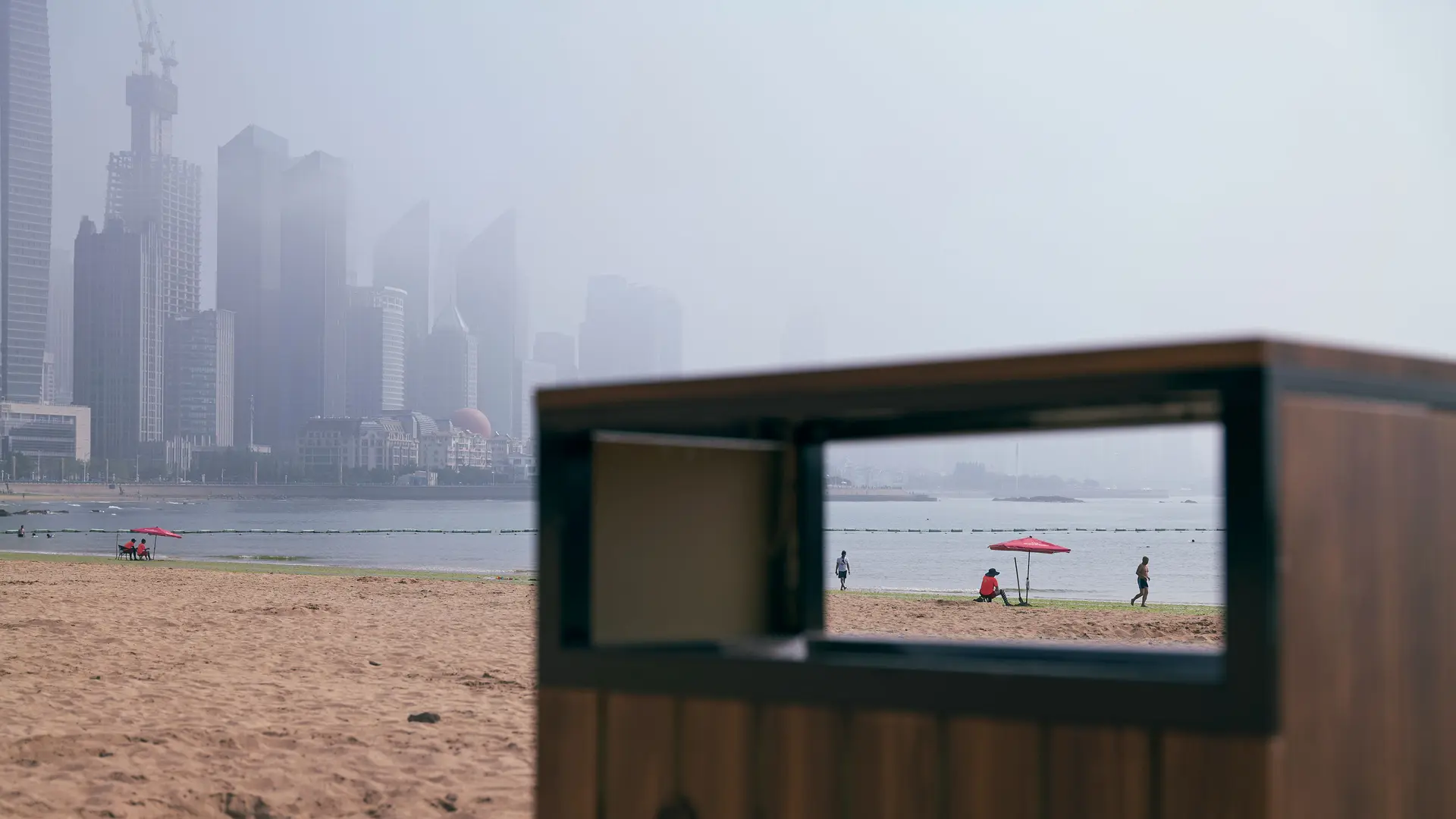 View through the rectangular opening of a garbage can on the beach with skyscrapers in the foggy background.