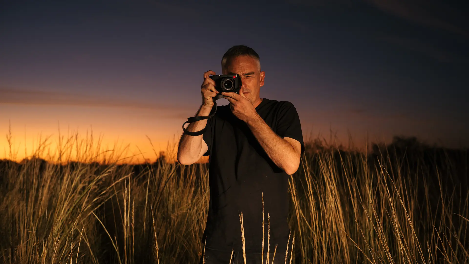 Person in a field with tall grass taking photos with a camera at sunset.