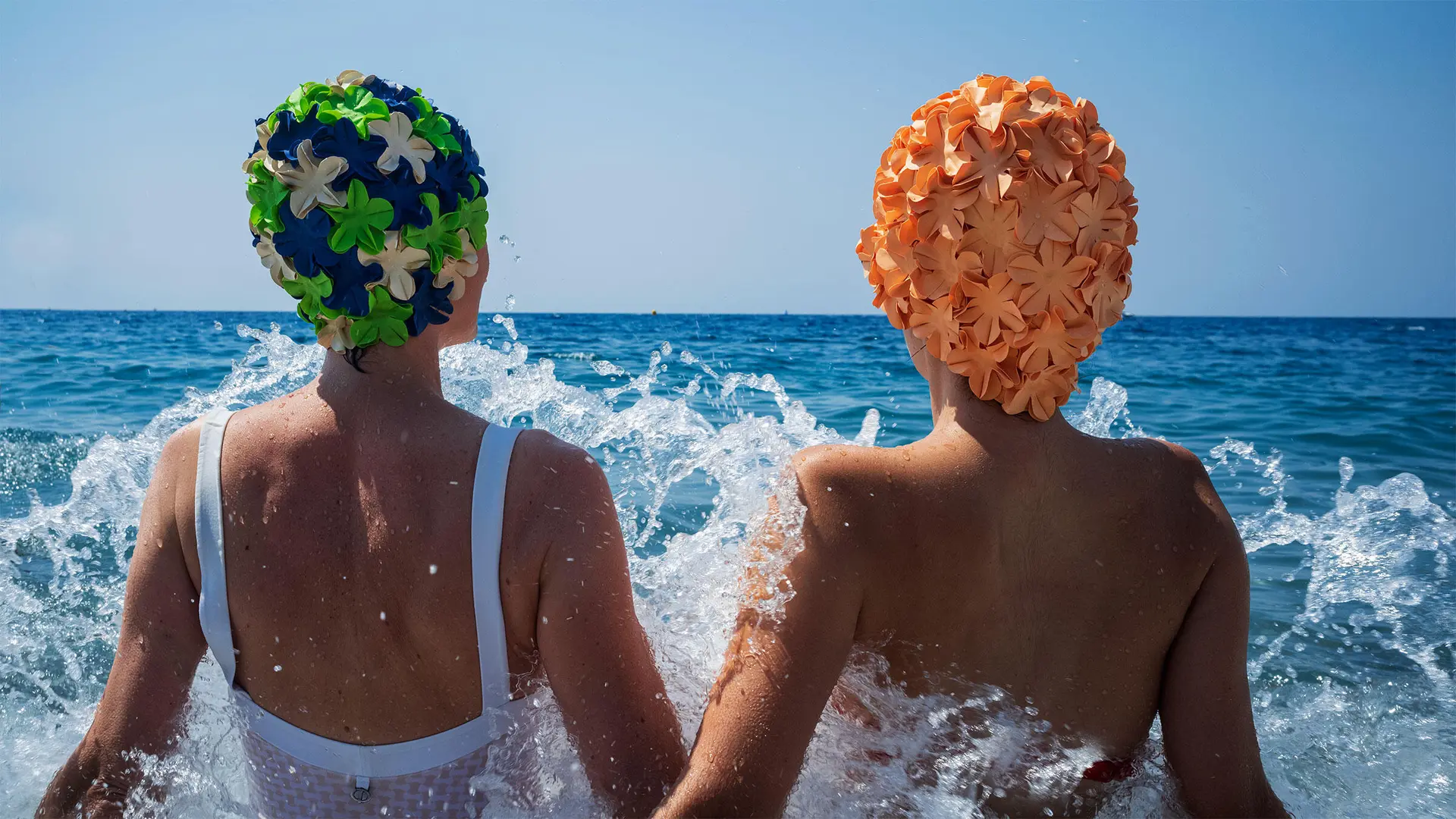 Two people in swimsuits and colorful bathing caps sitting in splashing water in the sea.