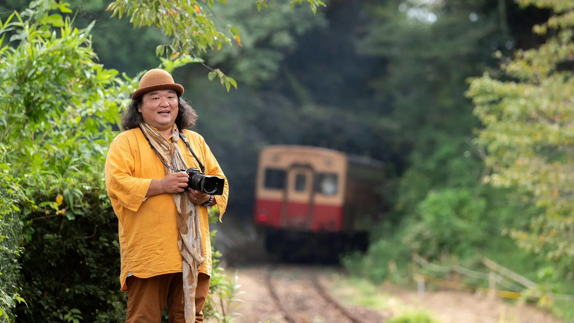 Man with camera stands beside railroad tracks and a train in background.