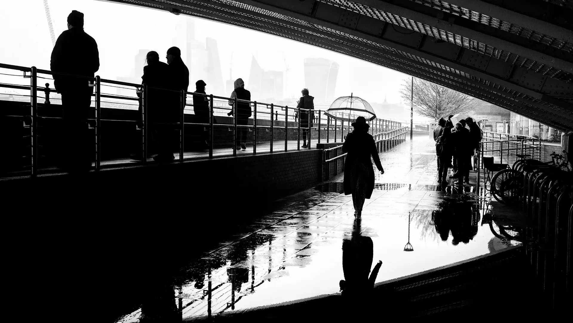 People walking under a bridge in the rain, silhouettes reflected in puddles.