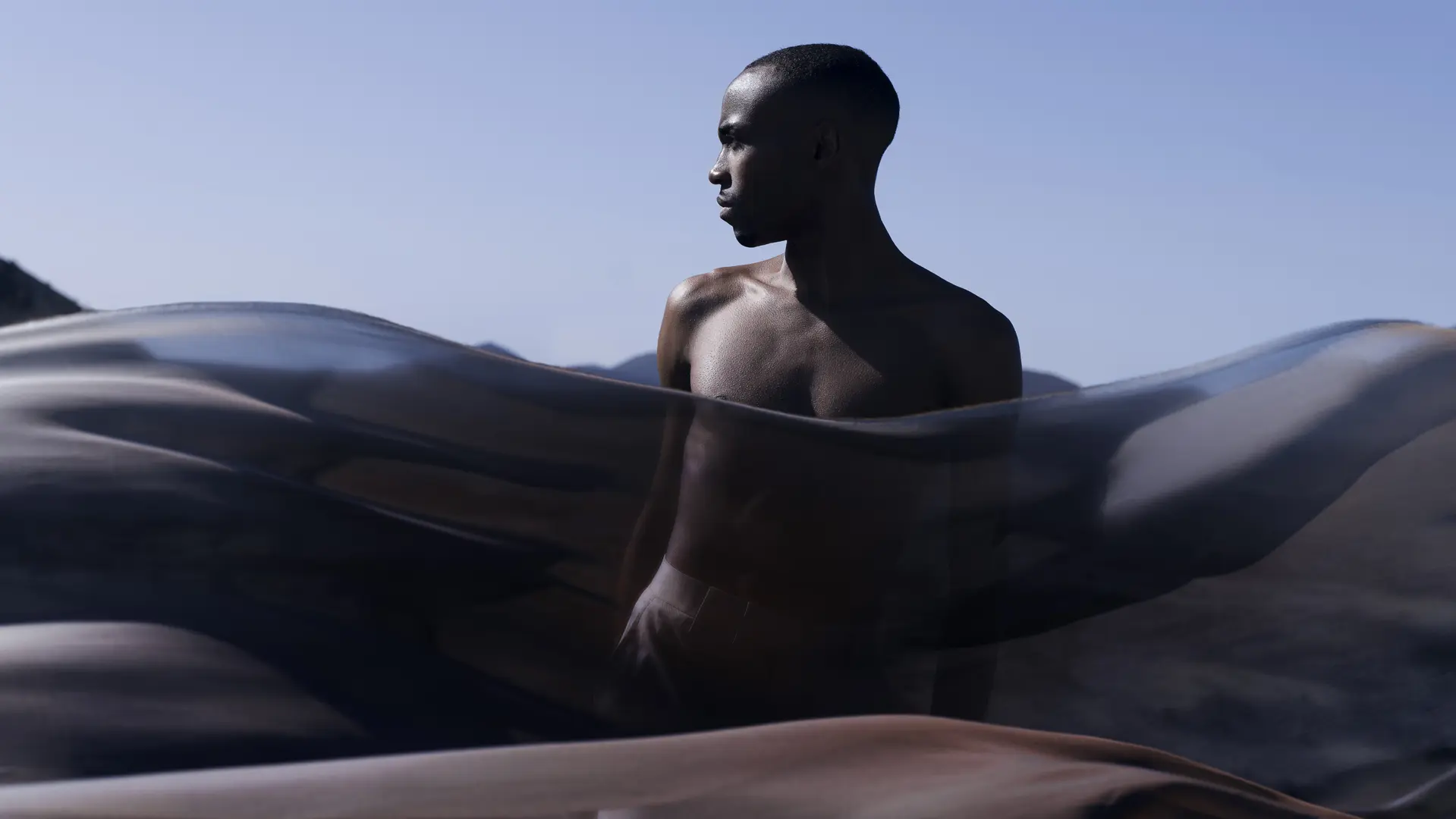 Man behind a transparent cloth looks to the side and light blue sky in the background.