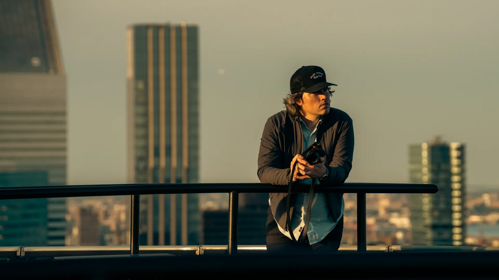 A man holding a camera leans against a railing with skyscrapers in the background.