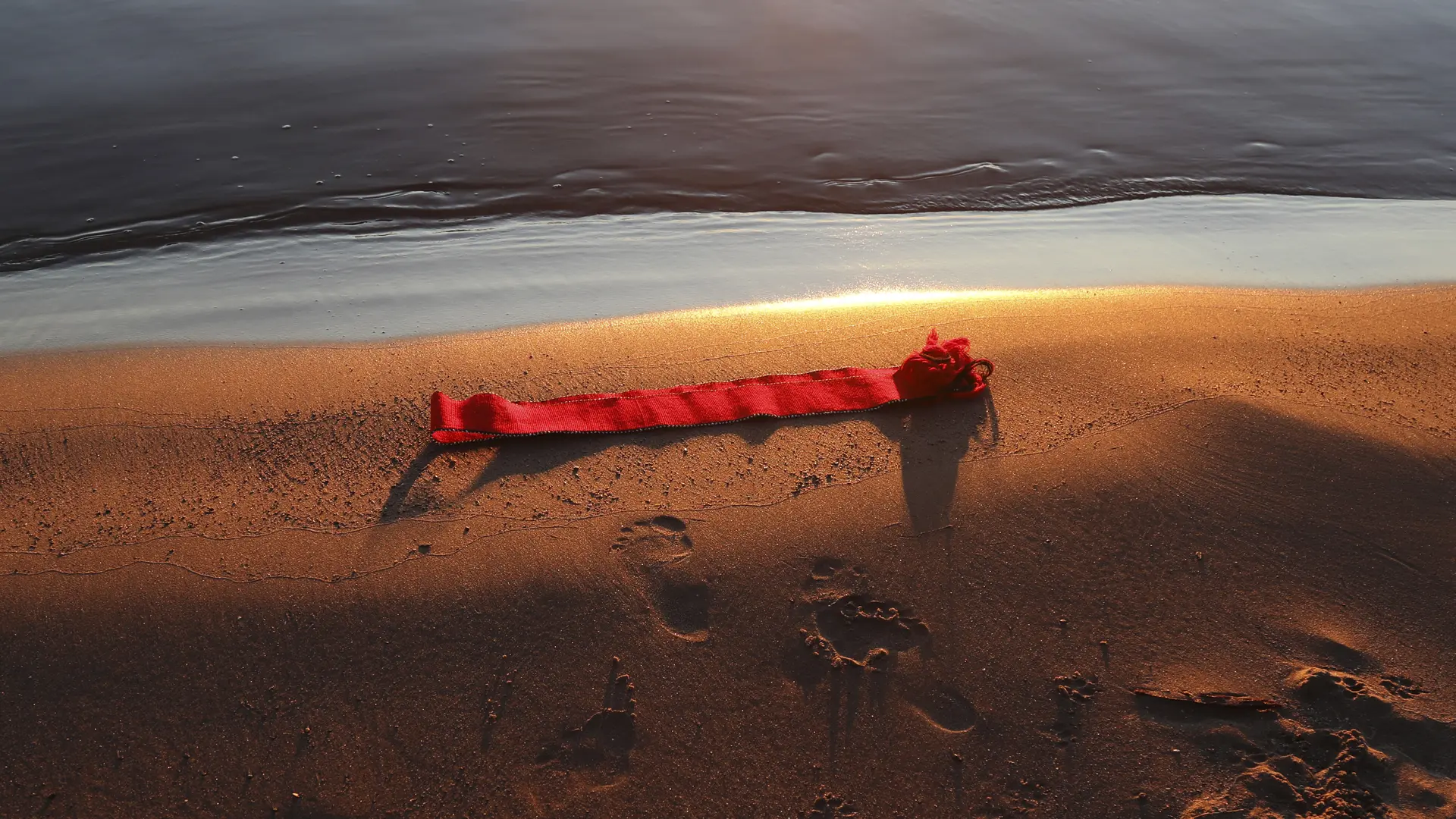 Red ribbon lying on the beach shore at sunset, footprints in the sand.