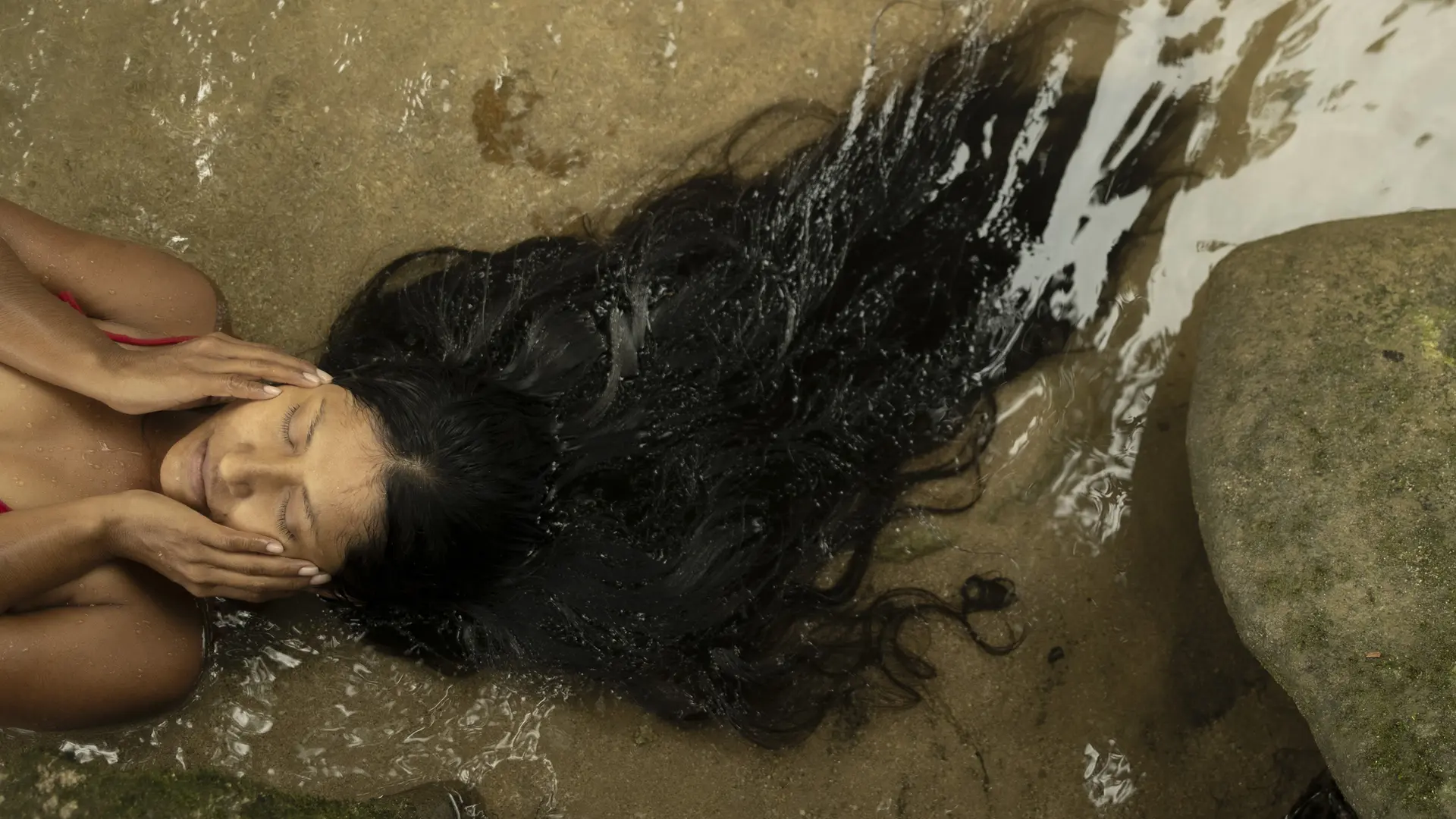 A woman is lying in the water, holding her face with both hands, her long hair floating in the water.