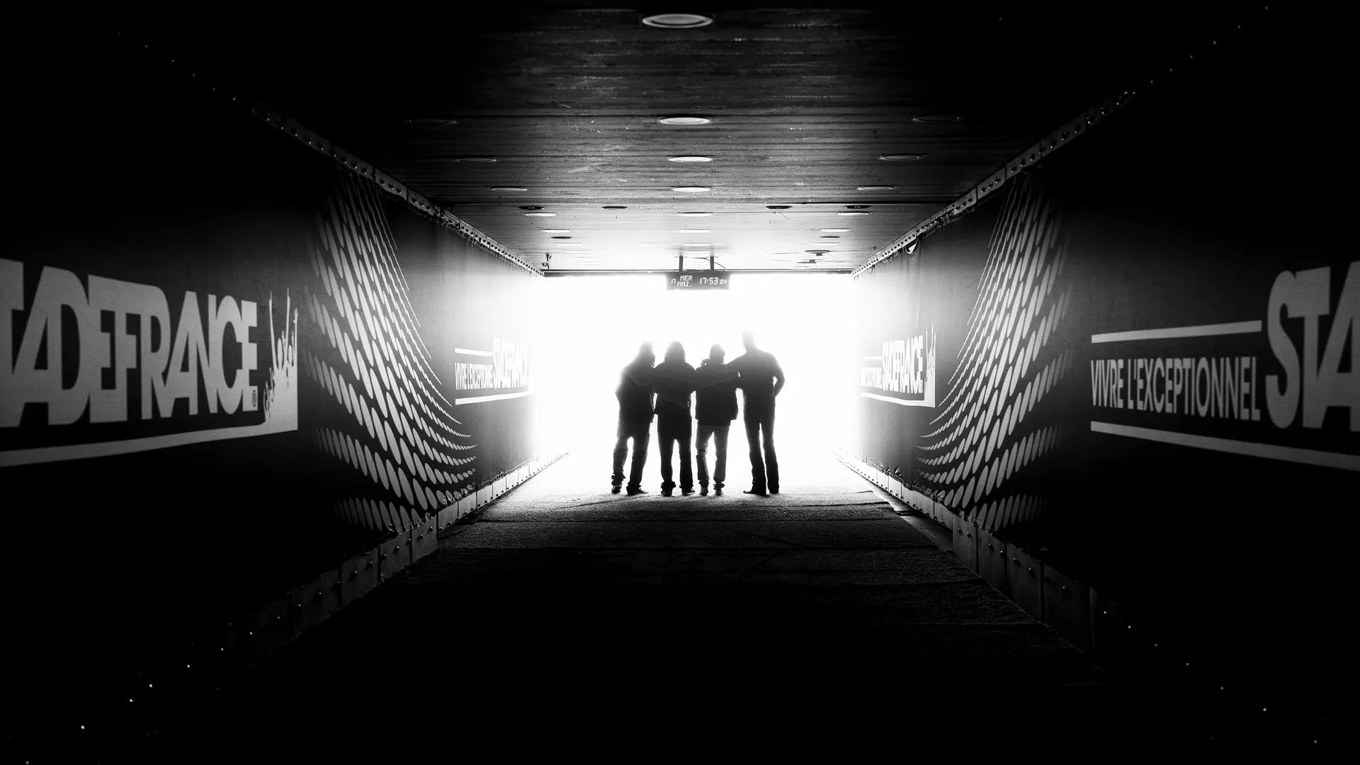 Four people are standing in a stadium tunnel with the sign "Stade France" in the light at the end of the corridor.