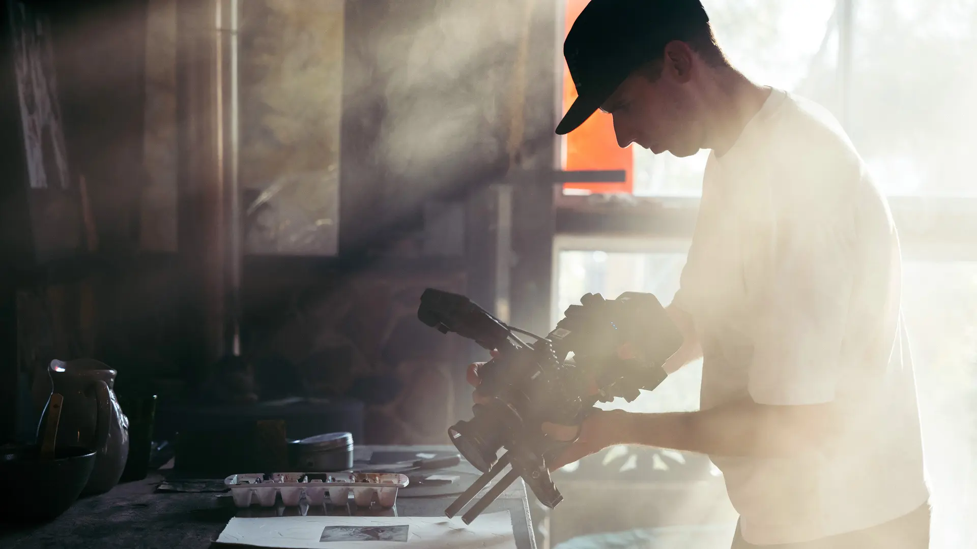 Person holds camera over a table with art materials, warm sunlight falls through window.