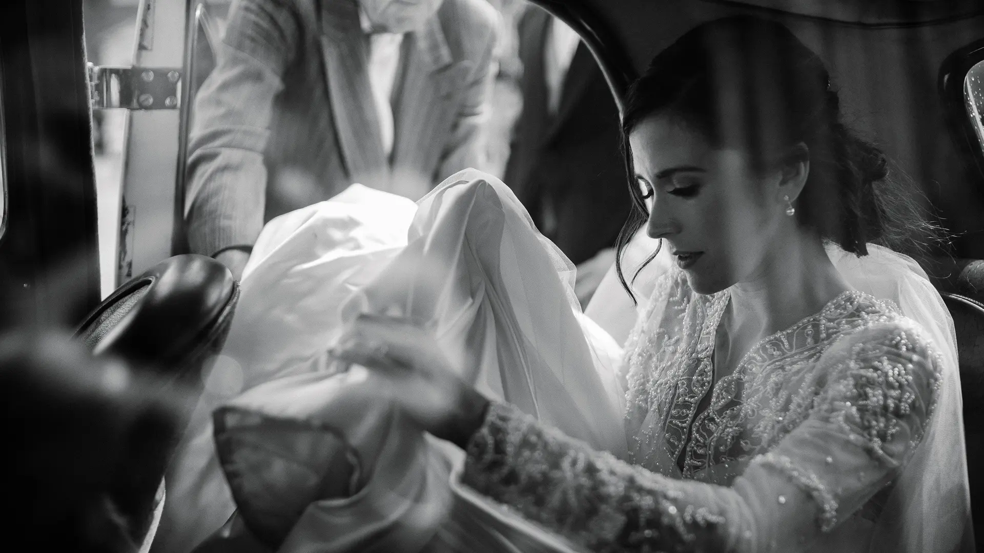 A bride sitting in a car holding her veil and a man standing next to the open car door.