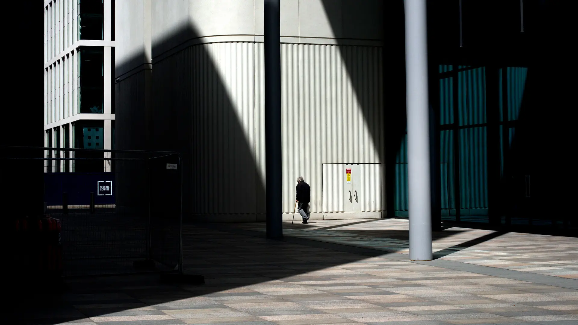 An old man walks in front of a building with shadows beside him.