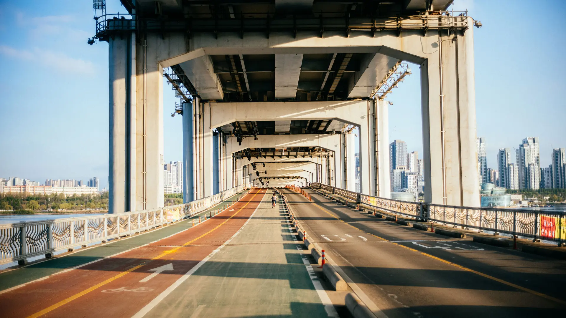 Single cyclist on a bridge with a separate cycle and pedestrian path and road, framed by tall city buildings in the background.