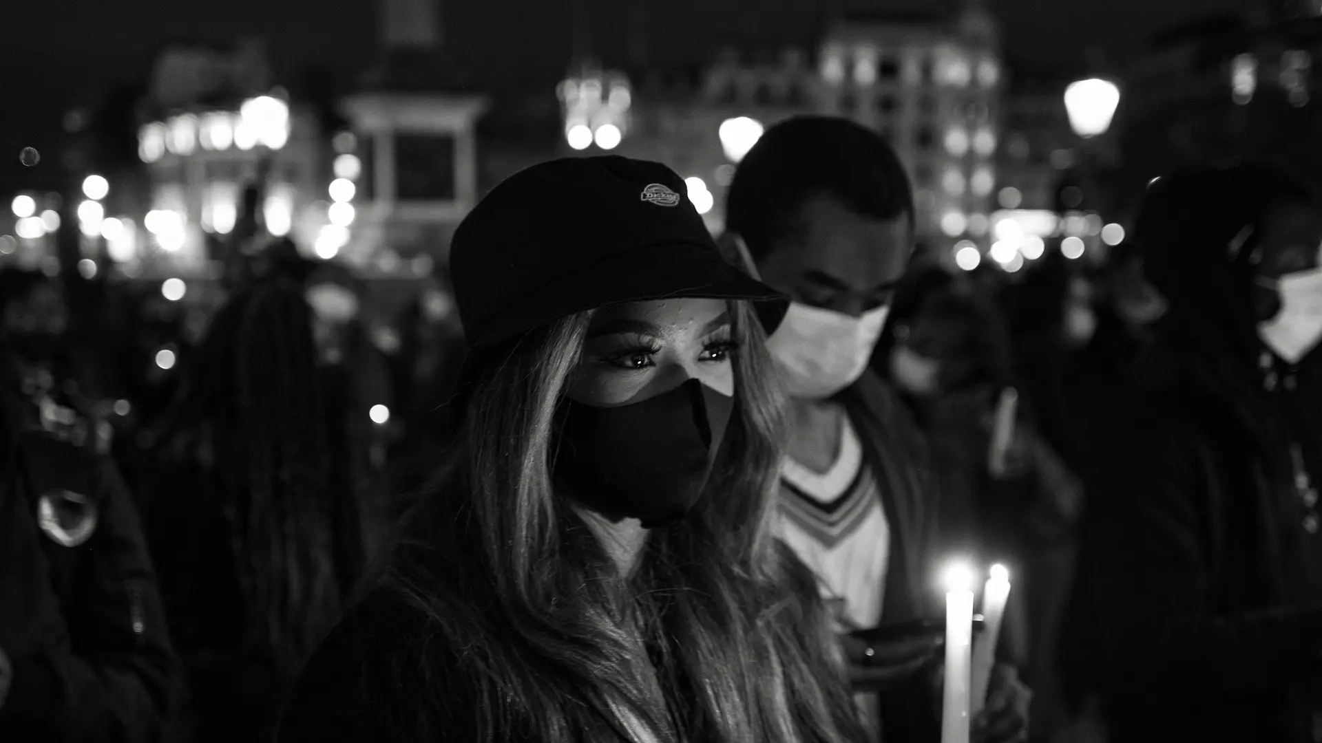 Close-up of a woman and man wearing masks and holding candles in a city at night.