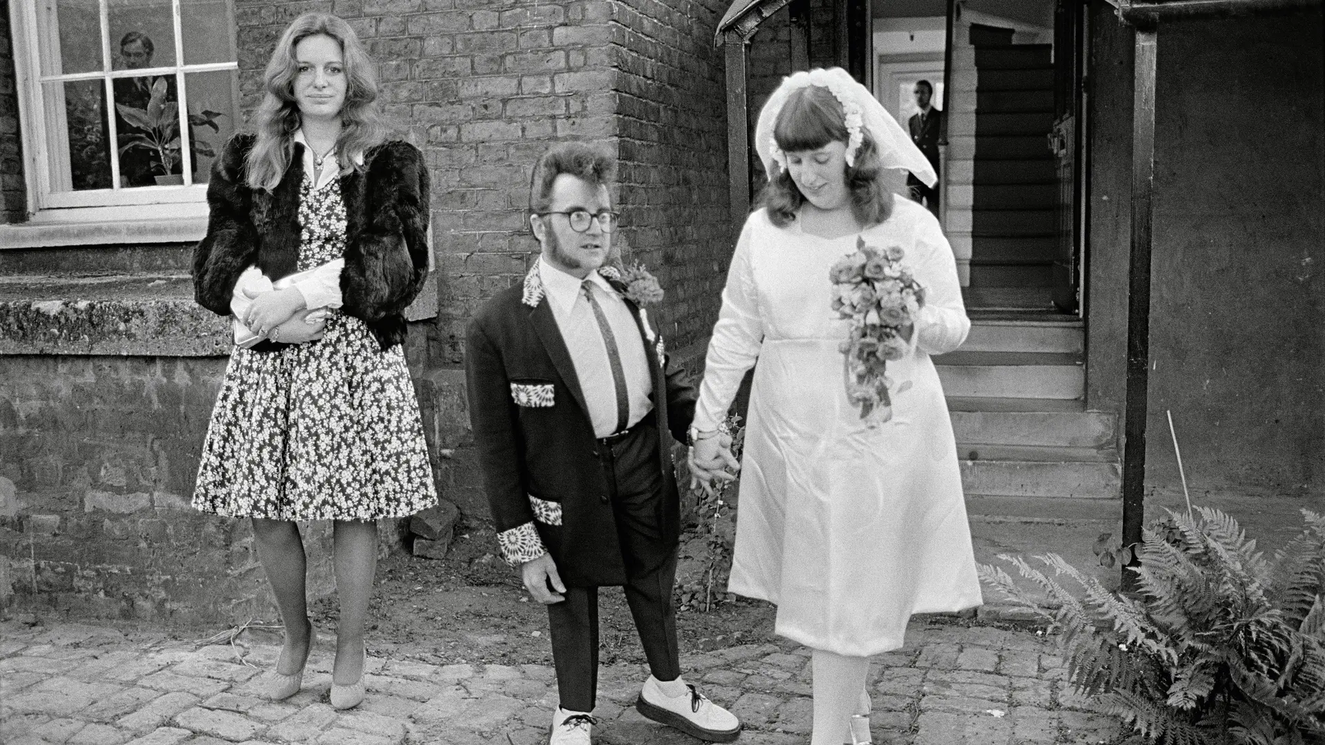 A wedding couple and a woman in a dress stand in front of a building.