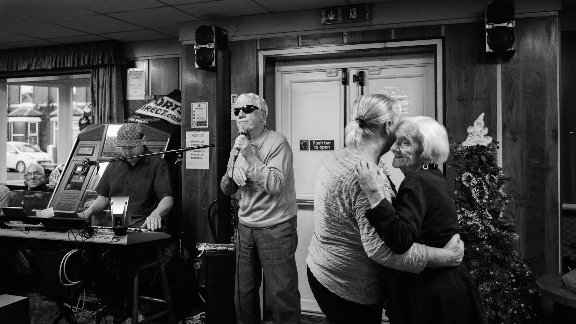 A man sings into a microphone while two older women dance and a man plays the keyboard in the background.