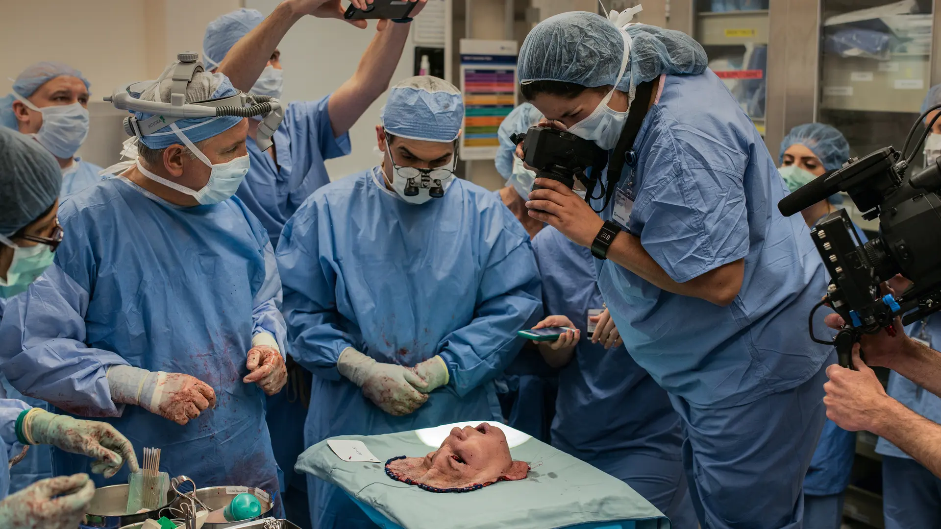 A group of people in surgical gowns look at a mask representing a face, and some take pictures of it.