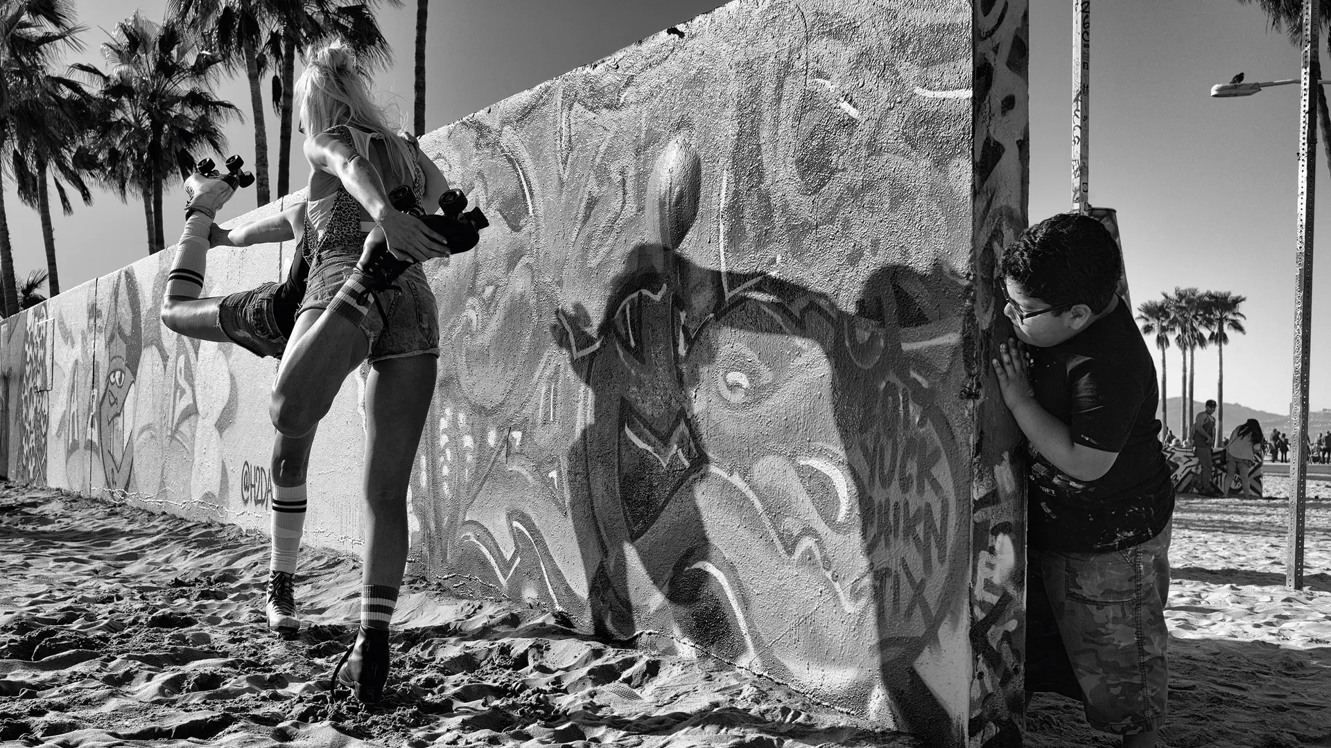 A boy looks behind a graffiti-painted wall at two women on roller skates warming up.