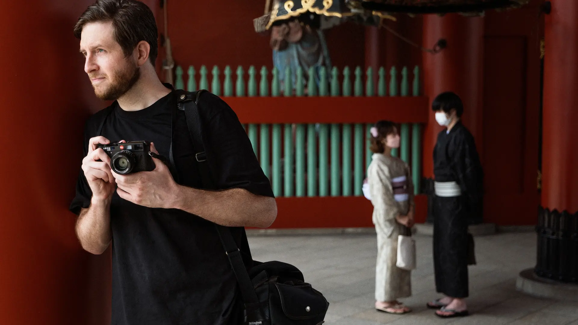 A photographer holds a camera while standing near traditional architecture, with two people in kimonos in the background.